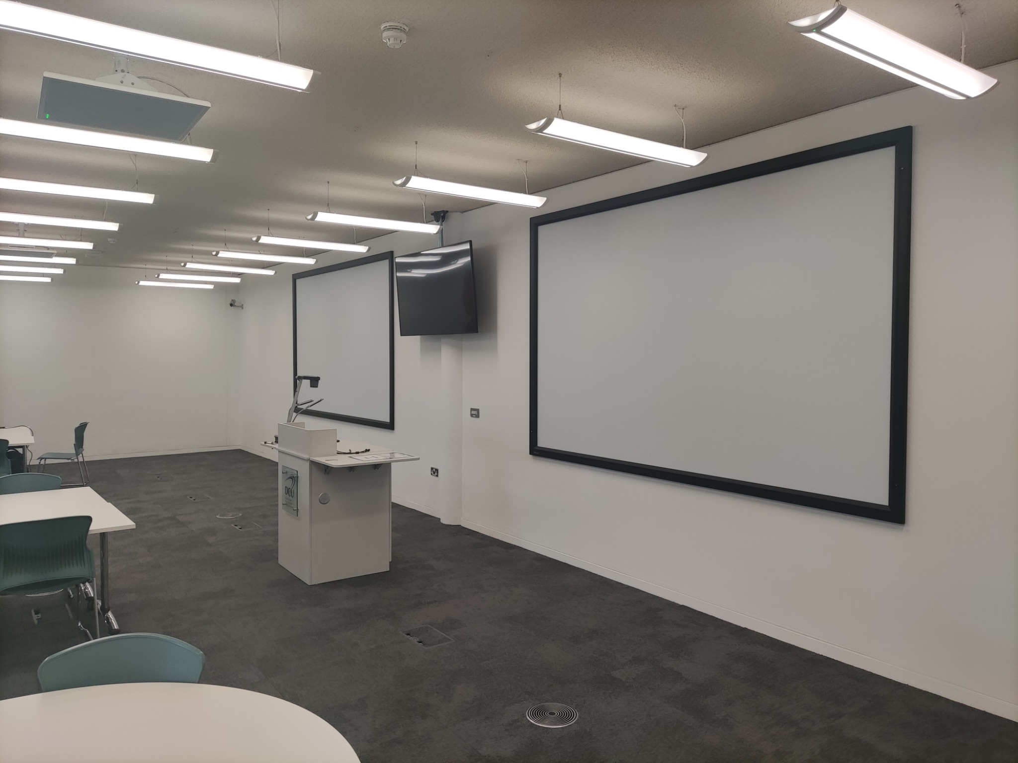 Conference room view from the back showing the presenter's lectern, two large projection screens on the front wall, and a wall-mounted monitor.