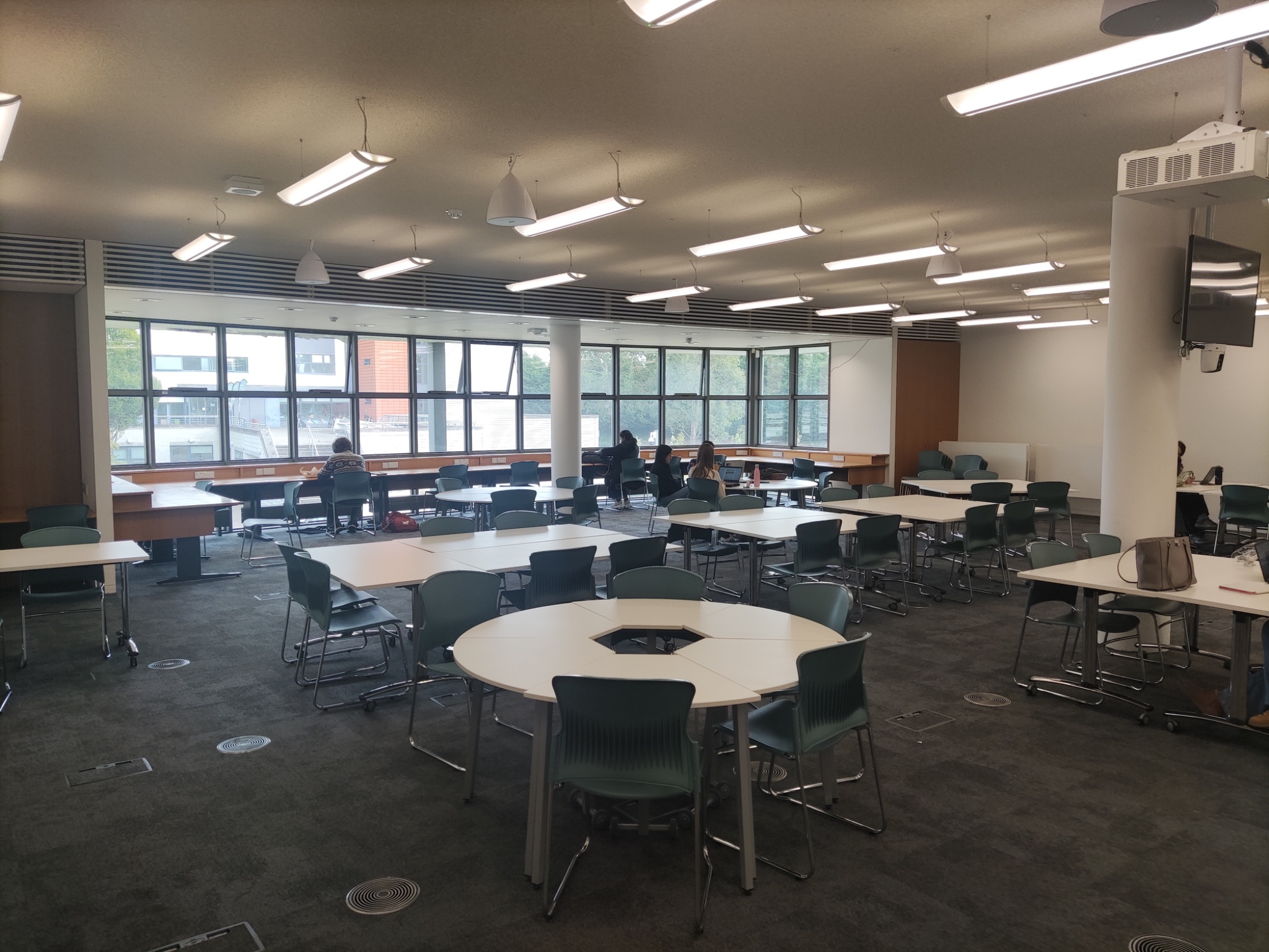 The Information Commons: a large open-plan room with round and rectangular tables, teal chairs, and large windows overlooking the campus.